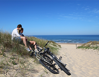 Par sidder i klitterne og ser ud over strand og hav med to cykler ved siden af sig